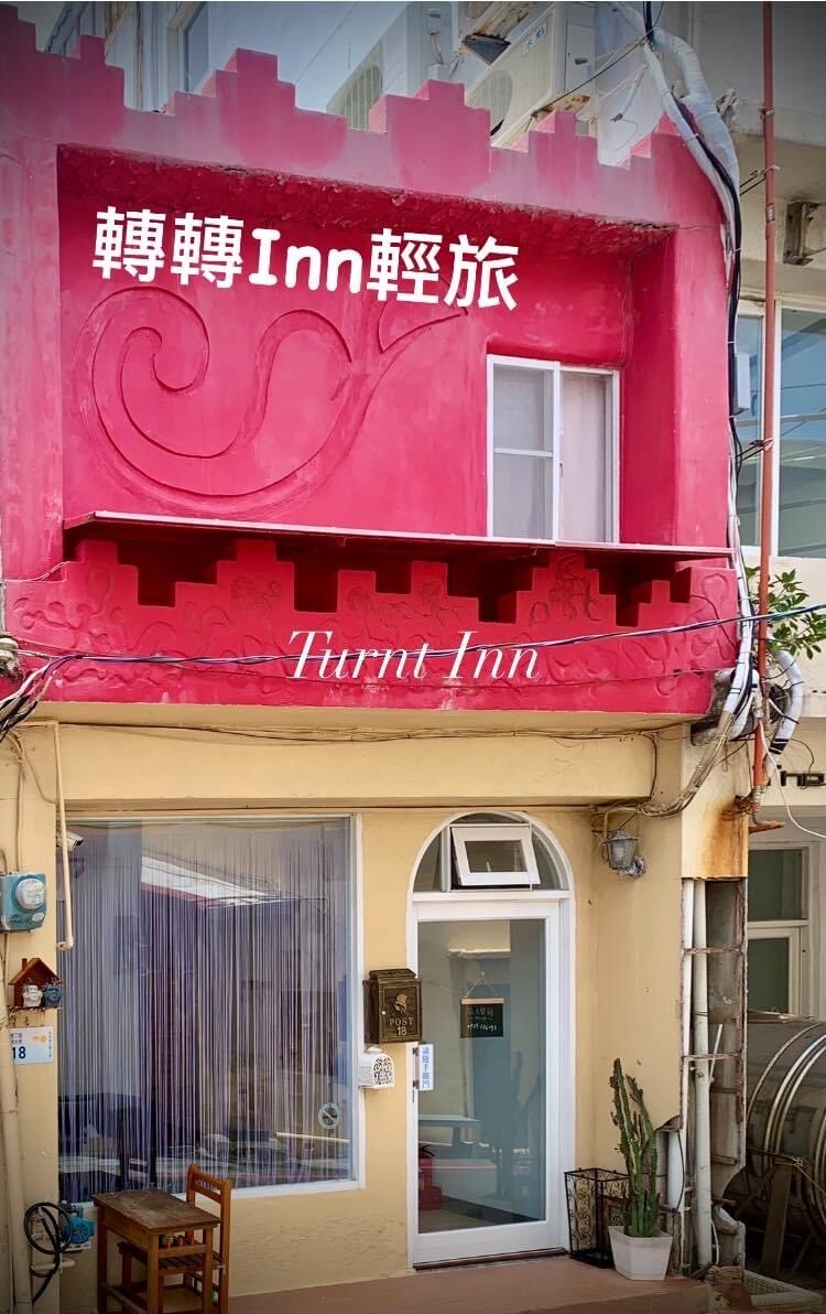 The exterior of the building features a vibrant pink facade, adorned with decorative elements. A white door with large glass panels provides access to the interior. A curtain partially covers the window, allowing for natural light, while text identifying the establishment is prominently displayed above the entrance.