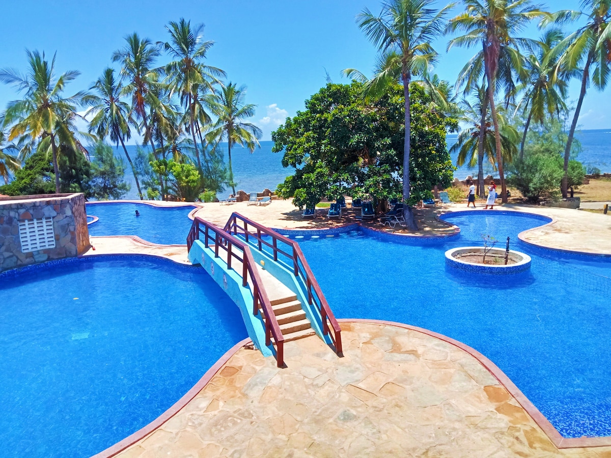 A large swimming pool featuring a wooden bridge spans across its surface. Surrounded by palm trees, the pool reflects the clear blue sky and offers views of the ocean in the background. Lounge chairs are visible along the poolside.
