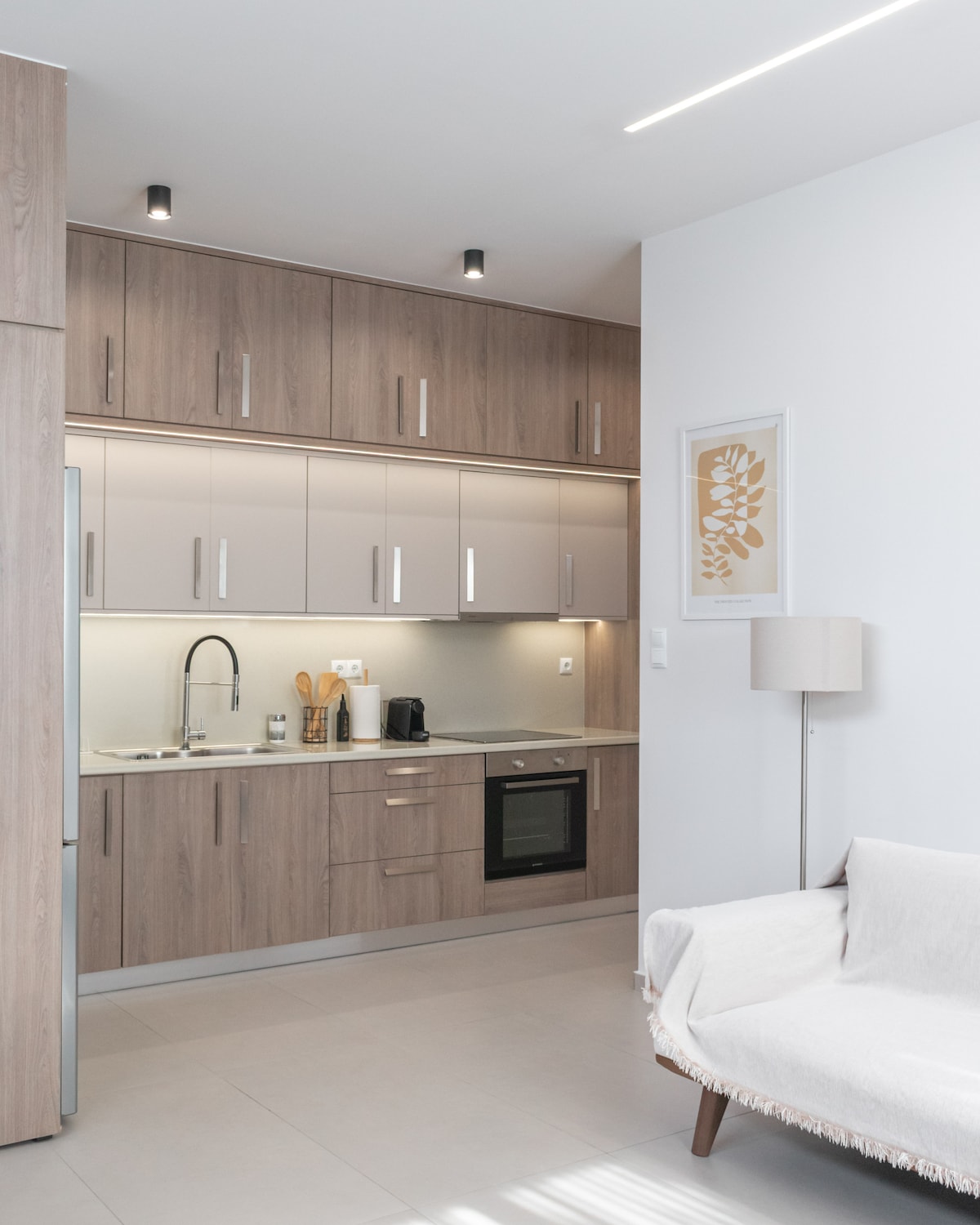 A modern kitchen is equipped with sleek wooden cabinetry, a stove, and a refrigerator. A sink is situated next to a countertop that holds various kitchen utensils. Soft lighting is provided by recessed fixtures, creating a bright and functional cooking space.
