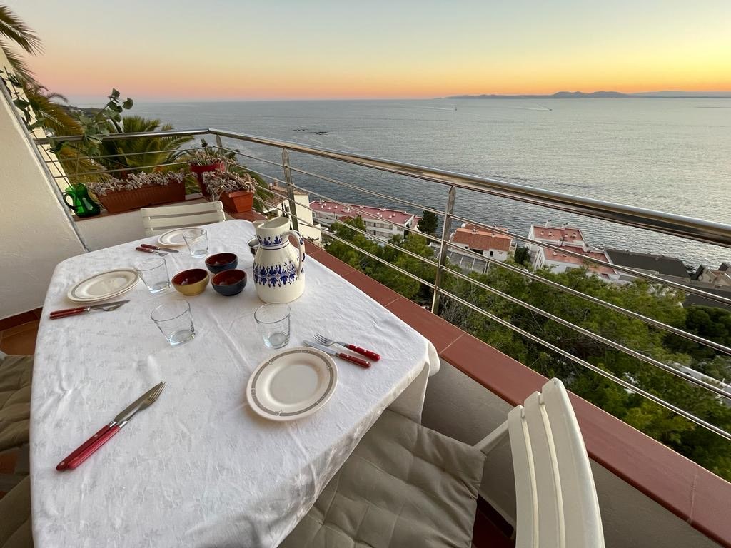 A terrace dining table is set for a meal, featuring white plates and cutlery. A scenic view of the sea and coastal landscape is visible in the background, complemented by a warm sunset casting soft colors across the sky.