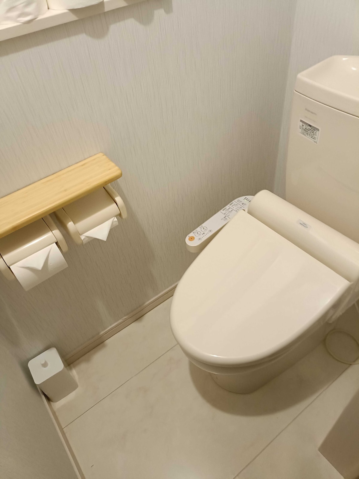 A modern toilet area features a sleek toilet and a wooden shelf holding two toilet paper rolls. The wall is adorned with light-colored paneling, and a small, discreet trash bin is positioned beside the toilet.