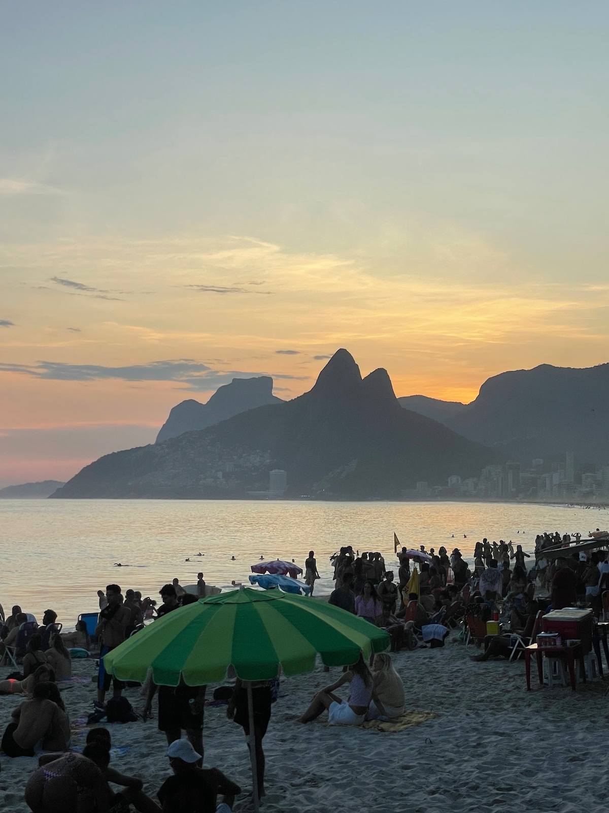 A lively beach scene is captured during sunset, with silhouettes of distant mountains forming a backdrop. Colorful umbrellas dot the sandy shore, and a crowd enjoys the water and warmth of the evening. Soft hues of orange and pink fill the sky, enhancing the vibrant atmosphere.