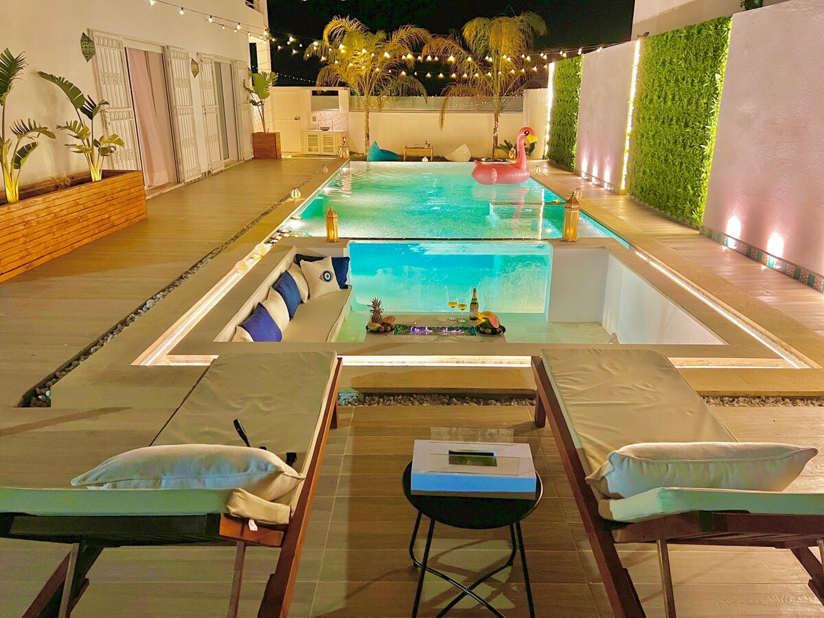 An outdoor area features a spacious infinity pool surrounded by loungers with plush cushions. Decorative elements include illuminated plants and soft lighting, enhancing the serene atmosphere. A hot tub is adjacent to the pool, with a vibrant pink float resting on the water.