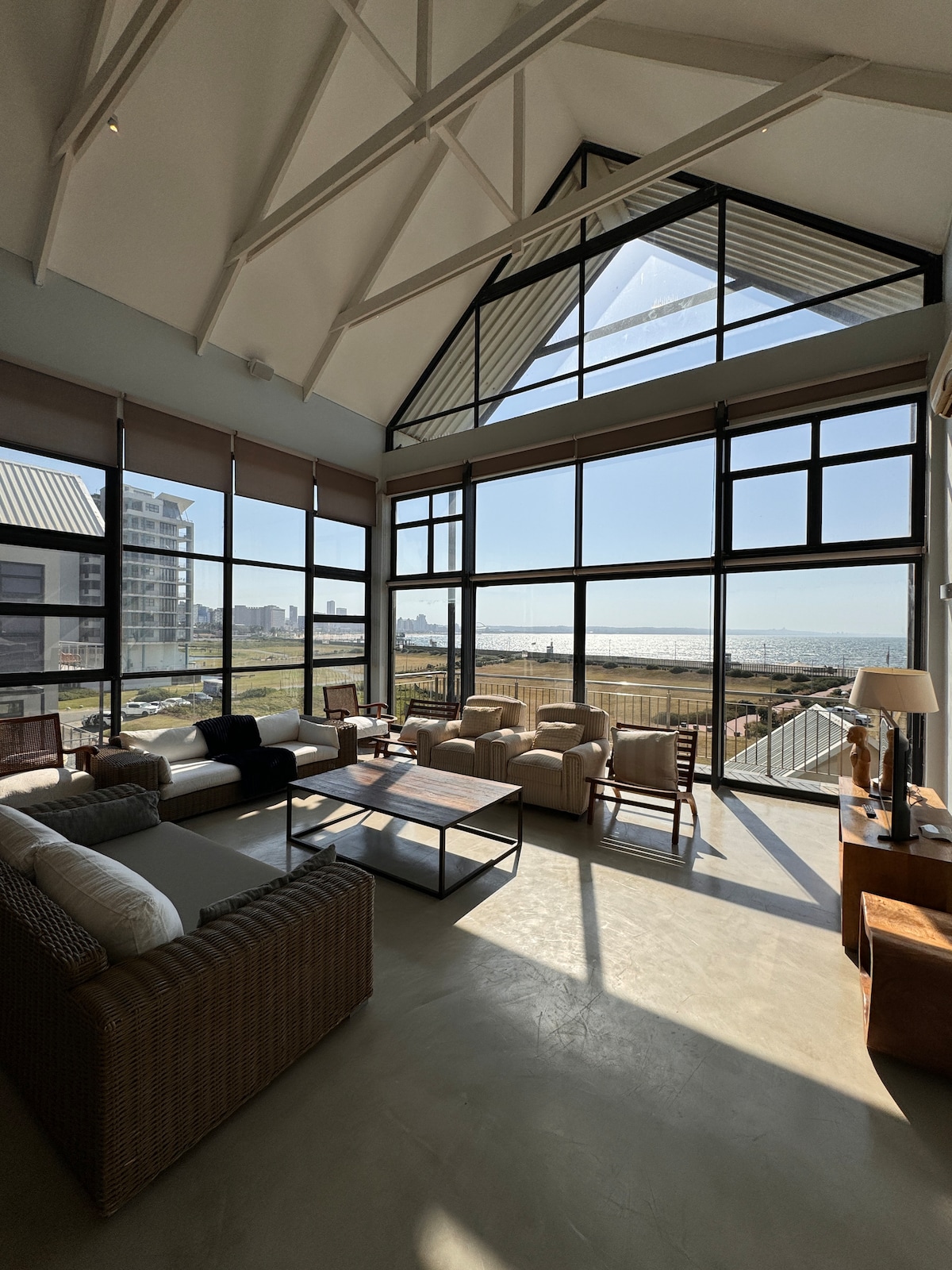 A bright and airy living area is featured with high sloped ceilings and large floor-to-ceiling windows, allowing natural light to flood the space. Comfortable seating is arranged around a central coffee table, providing a inviting spot for relaxation while offering expansive views of the surrounding area.
