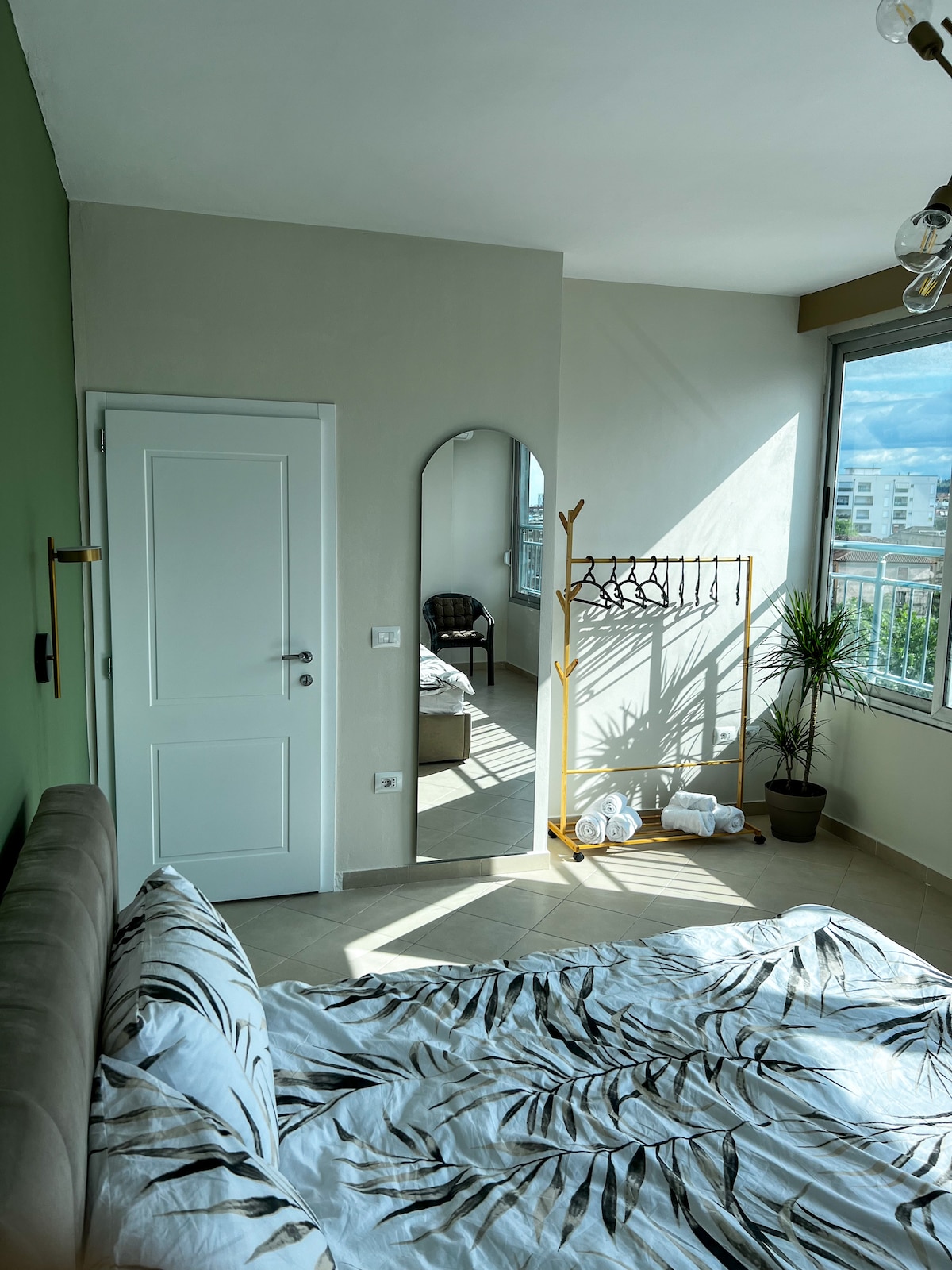 A bright bedroom features a large window allowing natural light to illuminate the space. A mirrored arch reflects the inviting atmosphere. A wooden rack holds towels, while a small potted plant adds a touch of greenery. The cozy bed is adorned with a tropical-patterned duvet.