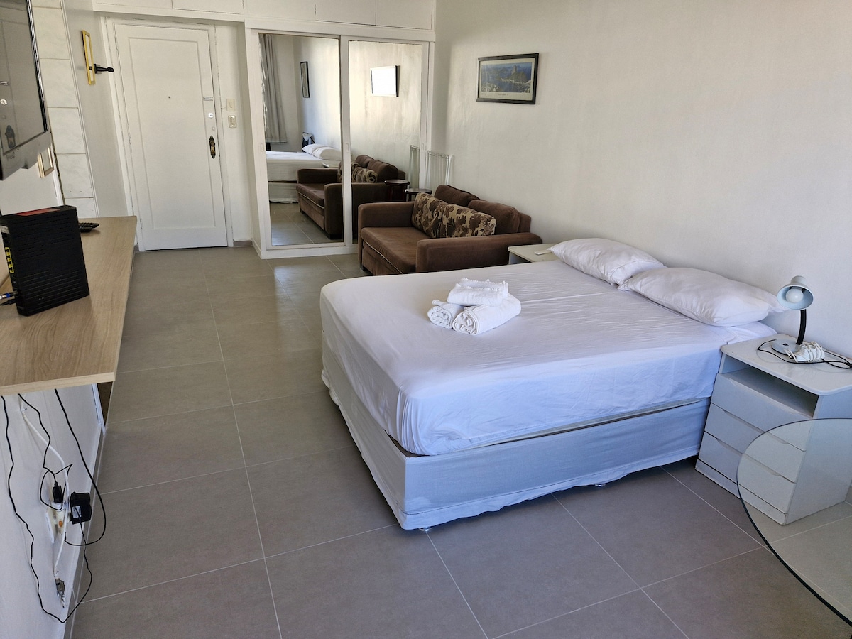 A comfortable sleeping area features a spacious bed with crisp white linens and neatly arranged towels on top. A brown sofa is located opposite the bed. A mirrored wardrobe reflects light and enhances the room's brightness. A bedside lamp rests on a small table beside the bed.