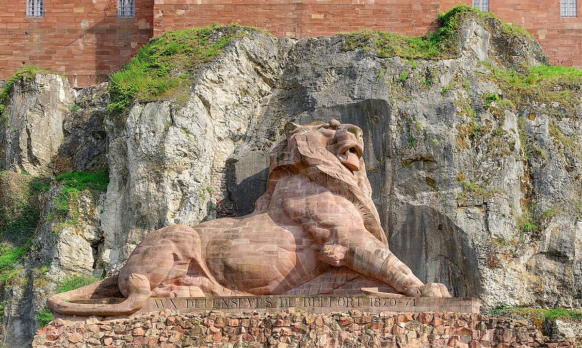 A large stone lion sculpture rests against a rocky backdrop, showcasing detailed features of the majestic creature. The lion is positioned on a stone pedestal, with a carved inscription visible in the foreground. Surrounding greenery adds a natural element to the scene.