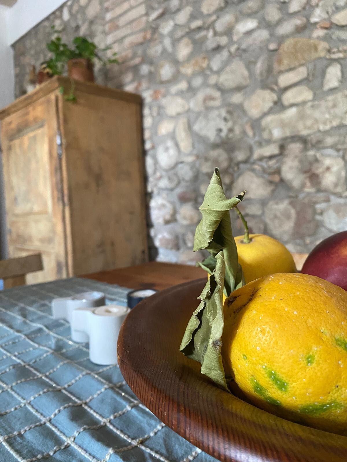 A wooden bowl filled with bright yellow lemons, green leaves, and red apples is positioned in the foreground. In the background, a rustic wooden cabinet is visible against a stone wall, while several white candles are arranged on a blue-patterned fabric.