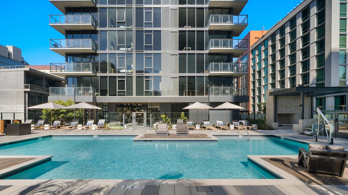 An outdoor pool is framed by a modern building with multiple balconies. Sun loungers are arranged around the pool, with large umbrellas providing shade. The surrounding area features well-maintained landscaping and ample open space for relaxation.