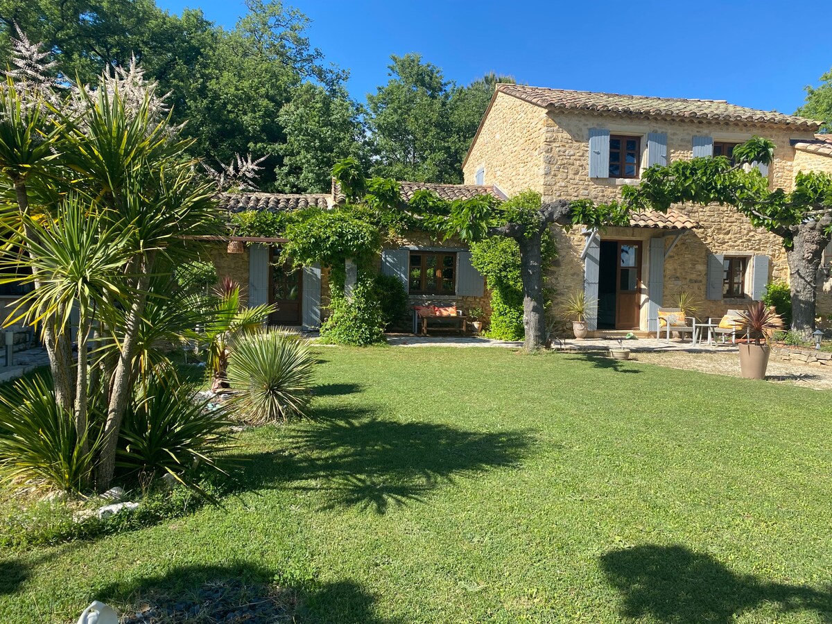 A charming stone house is set amidst a lush garden featuring well-maintained greenery and ornamental plants. The building's rustic facade is complemented by vines cascading over the walls. Outdoor seating areas are visible, inviting relaxation in the peaceful surroundings.