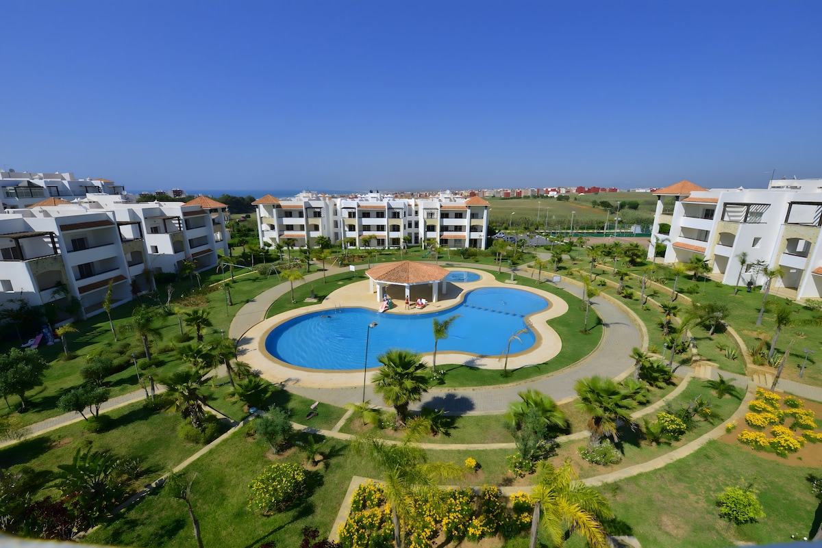 The image captures a spacious outdoor area featuring a large swimming pool surrounded by well-maintained gardens. Palm trees and colorful flower beds create a serene atmosphere. White buildings in a modern design are visible in the background, offering a view of the tranquil landscape.