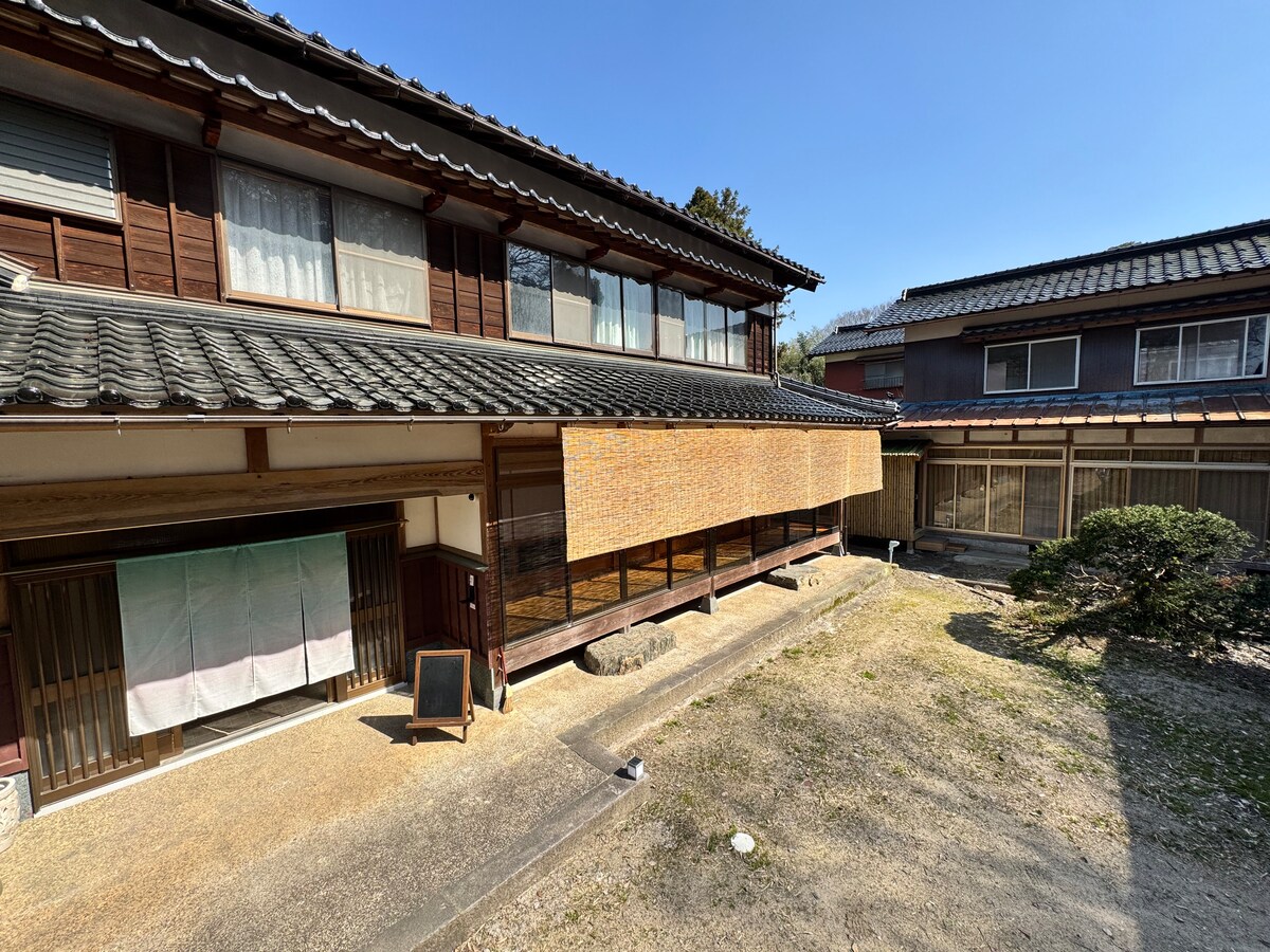 The exterior of a traditional guest house is visible, featuring a wooden facade and a cozy porch area with large windows. A sandy pathway leads to the entrance, and a black chalkboard sign stands ready for messages. Nearby, a garden area is framed by green shrubs.