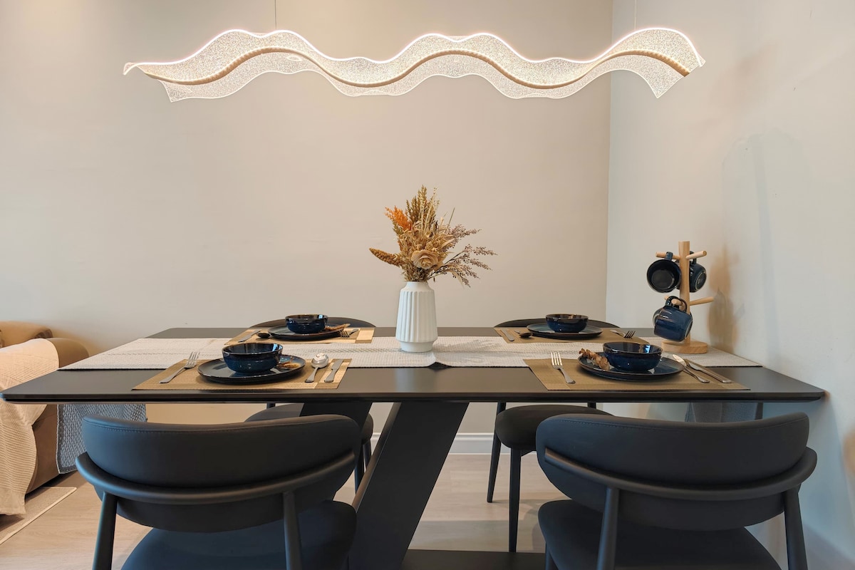 A modern dining table is set with four dark chairs, featuring elegant tableware including plates and cutlery. A central vase holds dried floral arrangements, while an artistic light fixture hangs above, adding a stylish touch to the dining area.