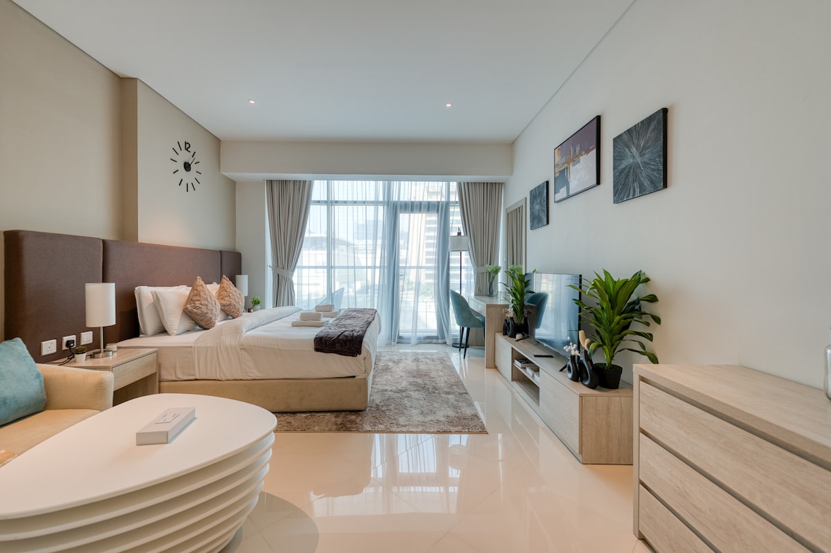 A spacious studio is adorned with modern furnishings, featuring a cozy seating area with a sofa and stylish coffee table. A queen-size bed is positioned against the wall, complemented by bedside lamps. Natural light fills the room through large windows, enhancing the serene ambiance.