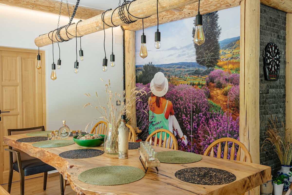 A spacious dining area features a large wooden table surrounded by multiple chairs. A vibrant mural of a natural landscape is displayed on the wall, adding color to the room. Soft lighting is provided by hanging bulbs, creating a welcoming ambiance.