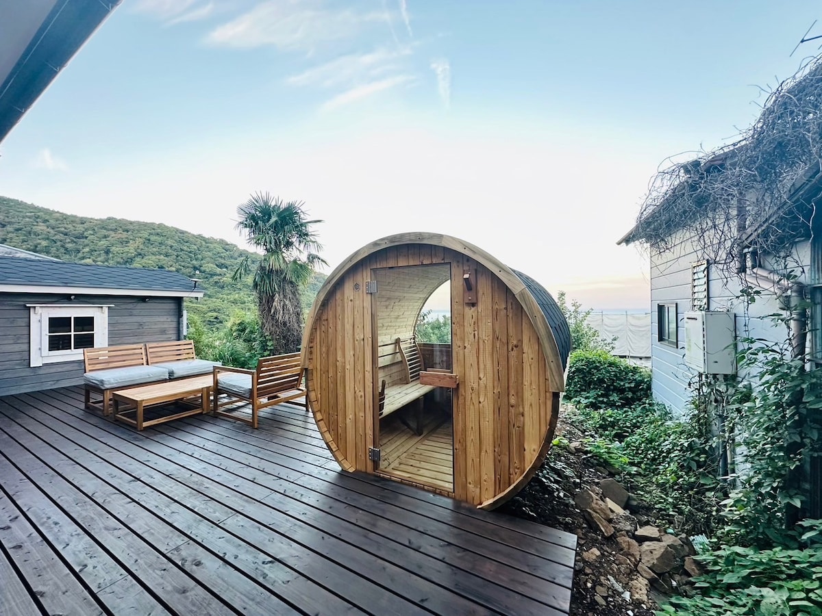 A wooden barrel sauna is positioned on a spacious deck, surrounded by lush greenery. Comfortable outdoor seating is arranged nearby, offering a relaxing space to enjoy views of the hills and ocean. The setting highlights a blend of nature and comfort.