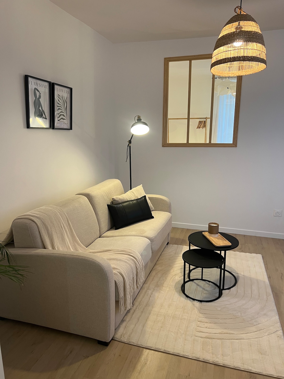 A soft beige sofa is positioned in a well-lit corner, complemented by a cozy throw and a black decorative pillow. A circular coffee table sits in front, adorned with a book and an elegant candle. Modern artwork frames the wall above the sofa, enhancing the inviting space.