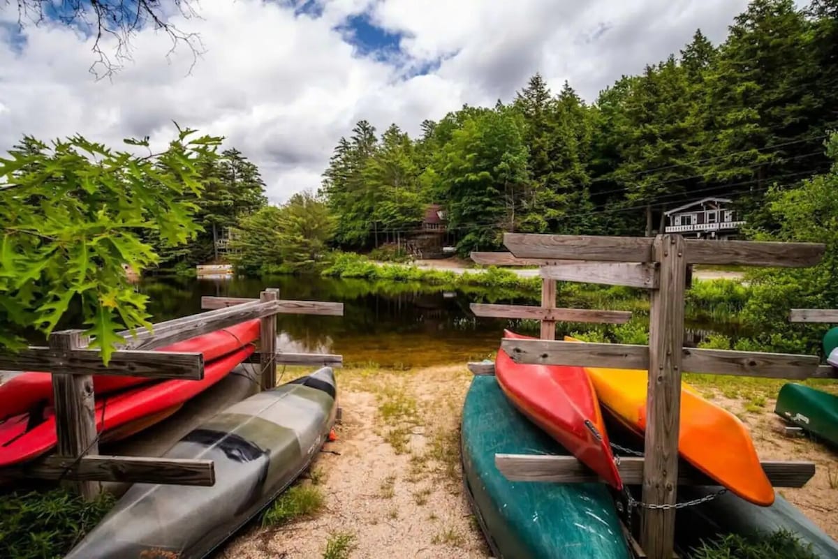 Colorful kayaks are stored near a serene body of water, surrounded by lush greenery. A wooden fence provides structure to the scene, while a rustic cabin is visible in the background. The clouds above lend a dynamic feel to the tranquil landscape.