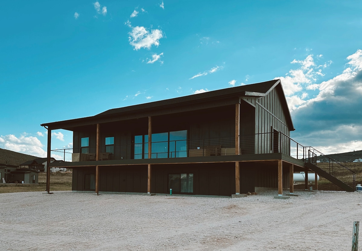 The cabin's exterior features a two-story structure with a modern design, large glass windows, and a covered porch. Natural light reflects off the windows, complementing the surrounding landscape. A gravel driveway leads to the entrance, showcasing ample parking space.