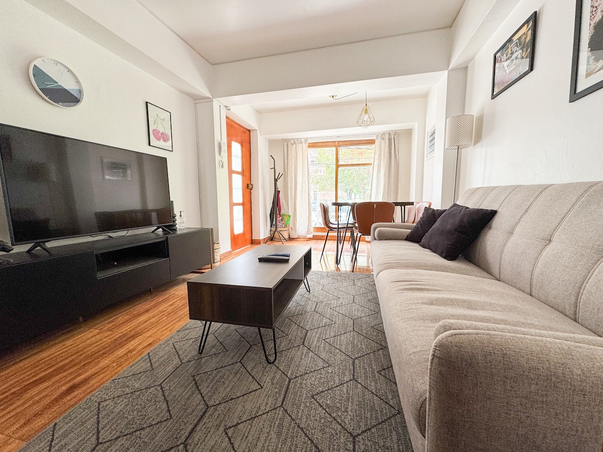 A well-lit living area features a comfortable sofa facing a large television. A coffee table with thin legs sits in front of the sofa on a geometric patterned rug. Natural light enters through sliding glass doors, complemented by a dining area in the background.