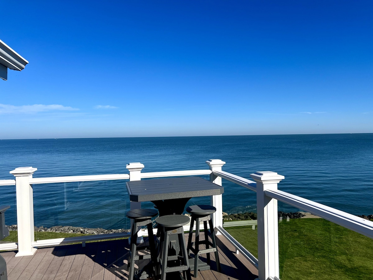 An outdoor deck features a table and four stools, overlooking expansive views of Lake Erie. The calm, clear water reflects the blue sky, creating a serene atmosphere. The deck is enclosed by white railing, enhancing the openness of the lakeside setting.