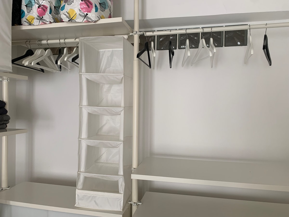 A modern closet features an organized layout with several hangers and a fabric storage unit with multiple compartments. Clean white shelving provides ample space for storing items, while towels are neatly stacked on a shelf above.