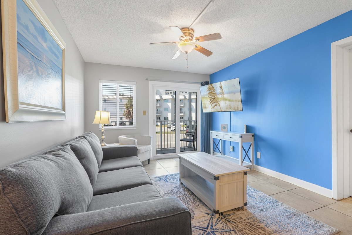 A bright living area features a gray sectional sofa and a coffee table atop a patterned rug. Light streams through the sliding glass doors, leading to a balcony with views. A flat-screen TV is mounted on the wall, complementing the blue accent wall.