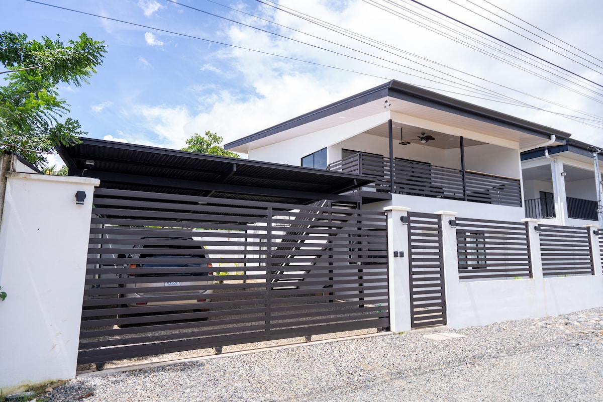 A modern apartment exterior is displayed, featuring a sleek black slatted fence and a covered parking area. A staircase leads to the upper level, enhancing accessibility. The vibrant sky provides a backdrop, complementing the architectural lines of the structure.