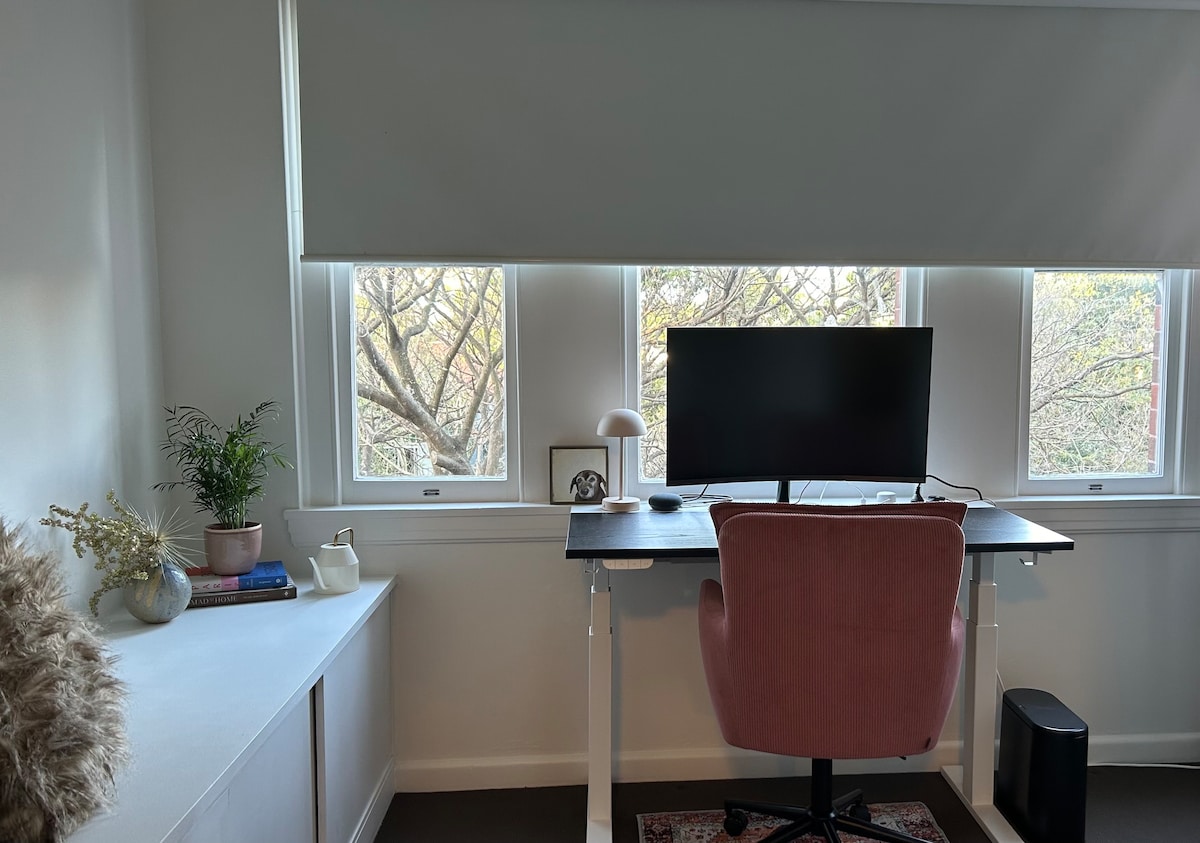 A private workspace features a modern adjustable desk, comfortable chair, and a large curved monitor. Natural light enters through multiple windows adorned with simple coverings. A small potted plant and decorative items are placed on the nearby shelf, providing a serene atmosphere.