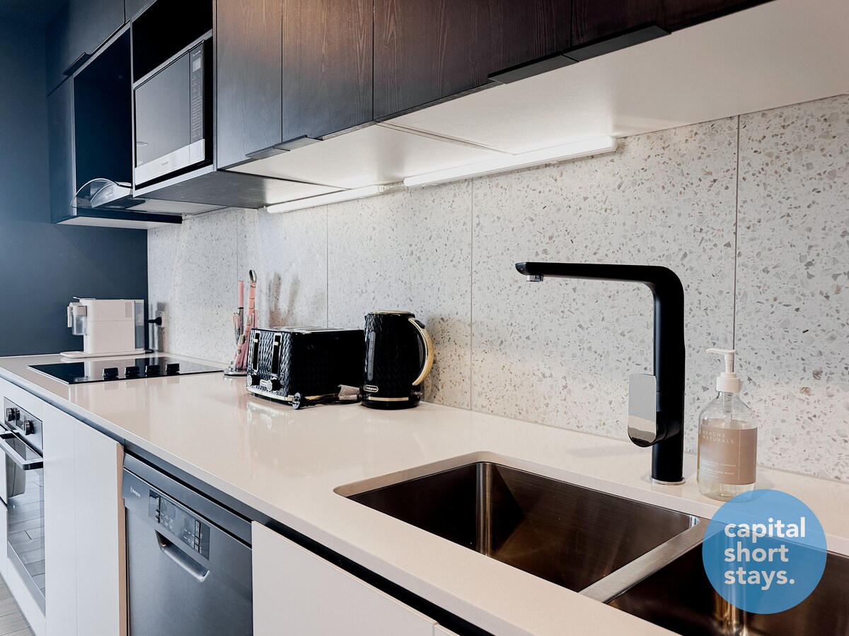 A modern kitchen is featured, equipped with high-end stainless steel appliances, including a dishwasher and microwave. Sleek stone countertops provide ample space for meal preparation, while under-cabinet lighting enhances visibility. A stylish sink and kitchen utensils are also visible, presenting an efficient and functional cooking area.