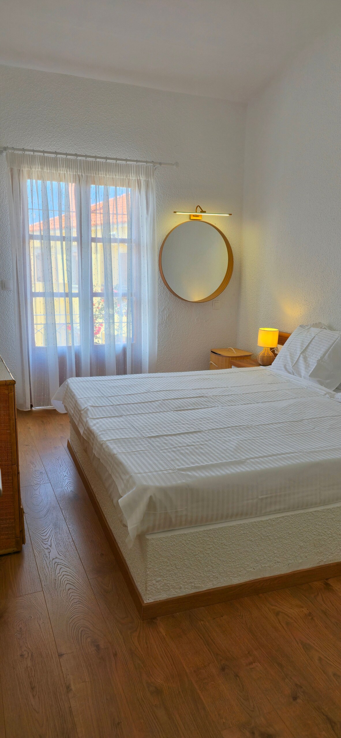 A bright bedroom features a spacious bed with crisp white linens, positioned near a large window that allows natural light to illuminate the space. A round mirror hangs above a bedside table, and a small lamp adds subtle warmth. Soft, light wood flooring completes the serene environment.