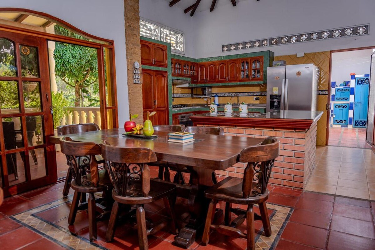A rustic wooden dining table with eight chairs is positioned centrally in a well-lit kitchen. Cabinets and countertops are visible in warm tones, while large glass doors open to a lush green area outside. Stainless steel appliances add a modern touch to the space.