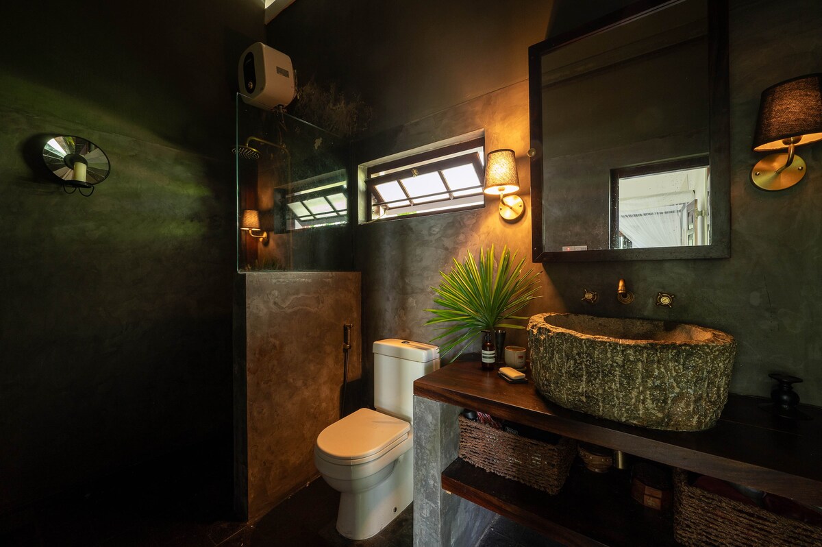A modern bathroom features earth-toned walls with a textured finish. A stone sink is placed on a wooden countertop, complemented by natural baskets underneath. Wall sconces provide gentle lighting, and a large mirror reflects the space. A glass shower enclosure is visible to one side.
