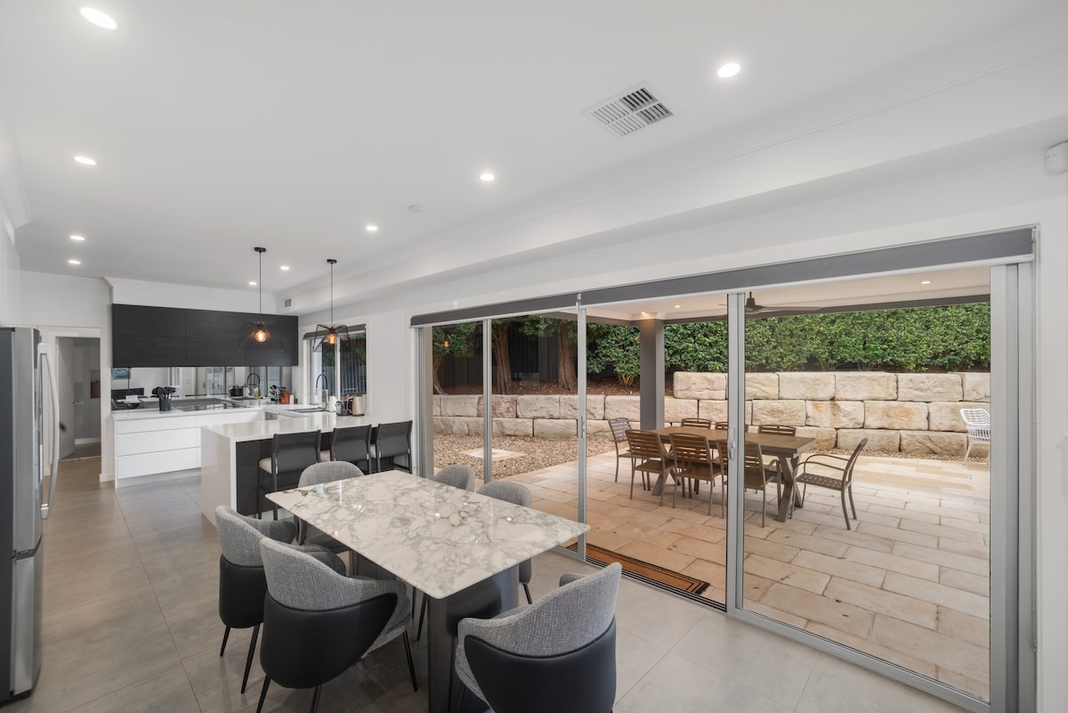 A modern kitchen and dining area features a large marble table surrounded by plush grey chairs. Sliding glass doors open to an outdoor patio with stone walls and additional dining options, enhancing the connection between indoor and outdoor spaces.