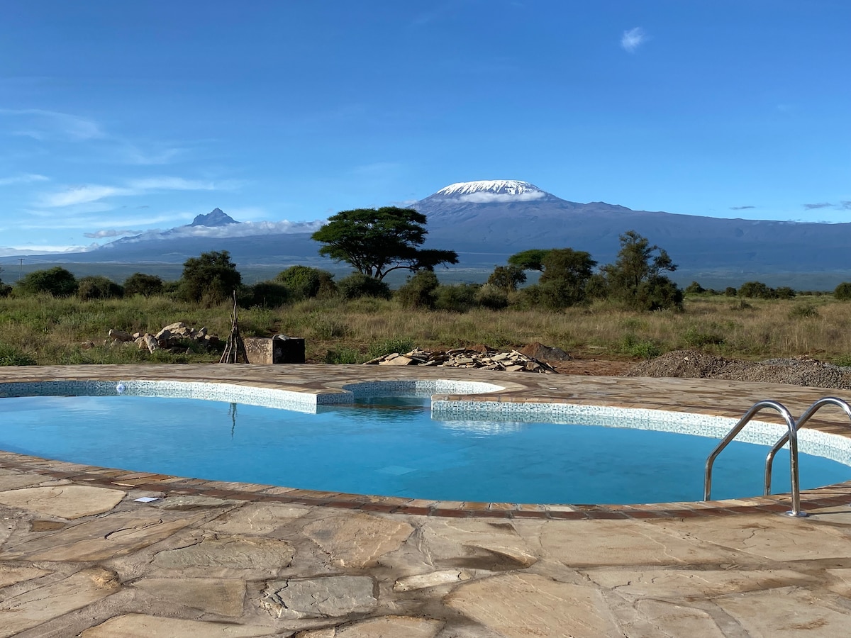 A clear blue swimming pool is surrounded by a stone patio, with a scenic view of snow-capped Mt. Kilimanjaro in the background. Lush green foliage and open grasslands can be seen, creating a peaceful atmosphere for relaxation.