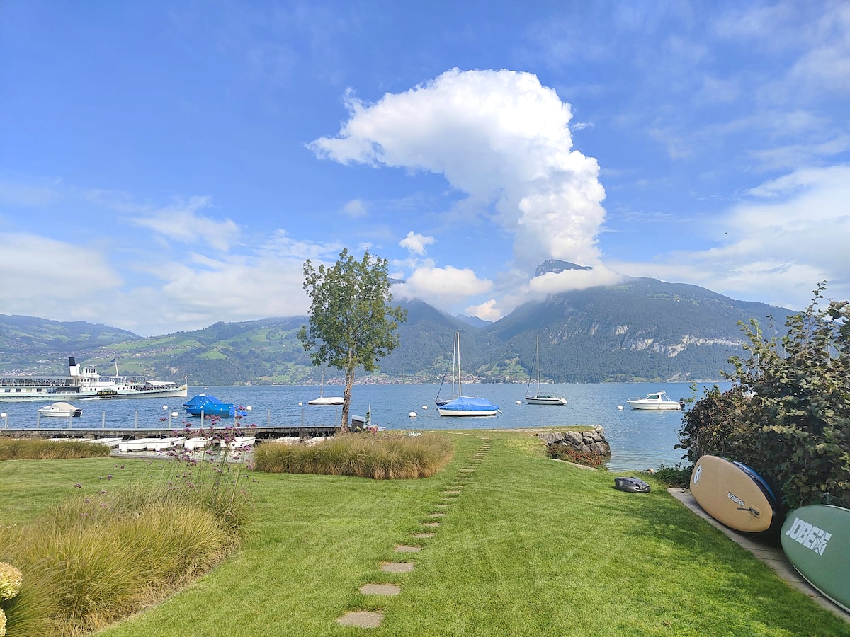 A scenic view of a serene lake setting is displayed, featuring sailboats on the water and lush green hills in the background. A well-maintained lawn with walking paths leads towards the water, while a paddleboard rests on the grass, inviting leisurely outdoor activities.