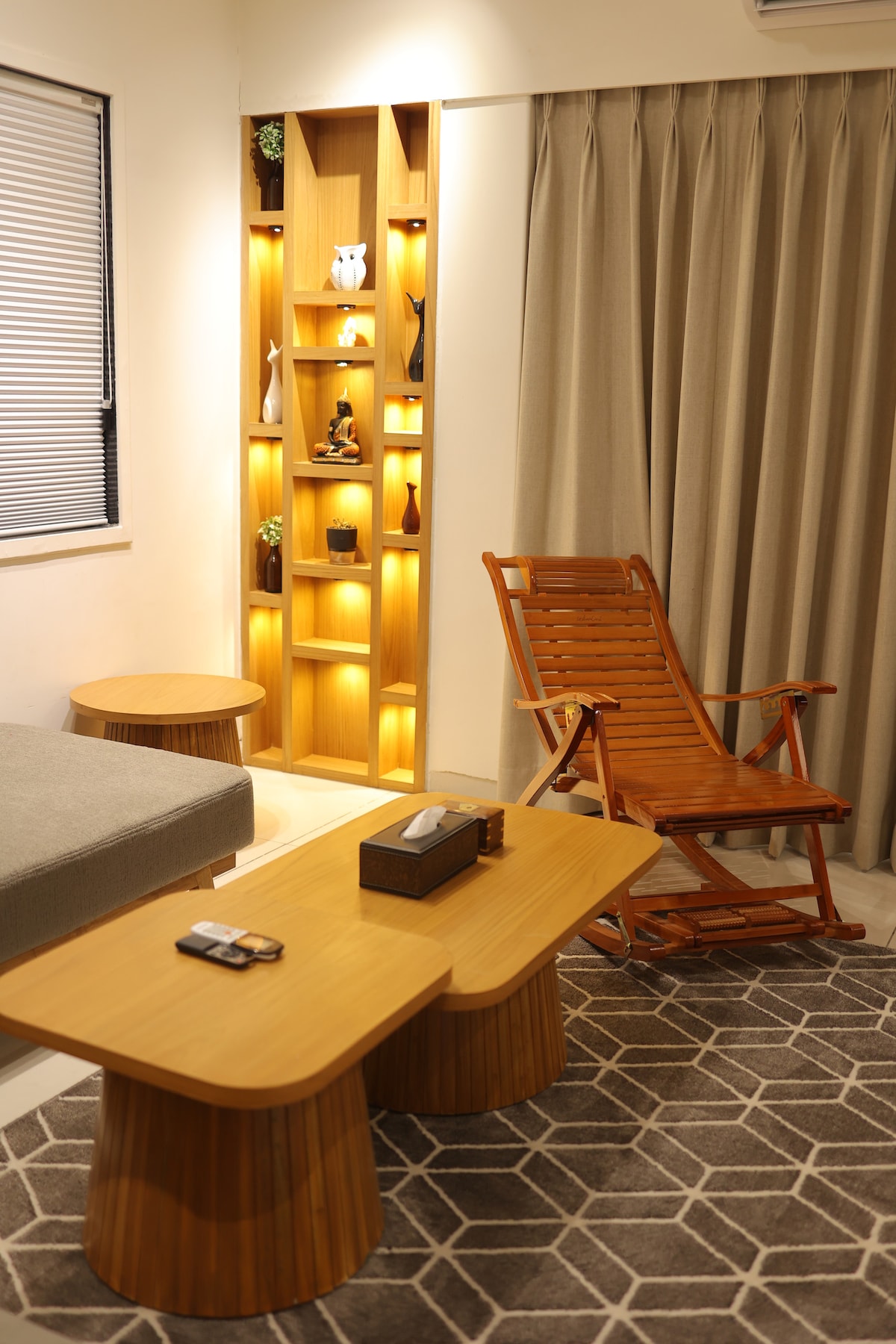 A cozy living area is highlighted by warm ambient lighting from built-in shelves displaying decorative items. A wooden rocking chair adds a touch of comfort, complemented by round coffee tables on a geometric-patterned rug beneath. Soft beige curtains frame the window, enhancing the serene atmosphere.