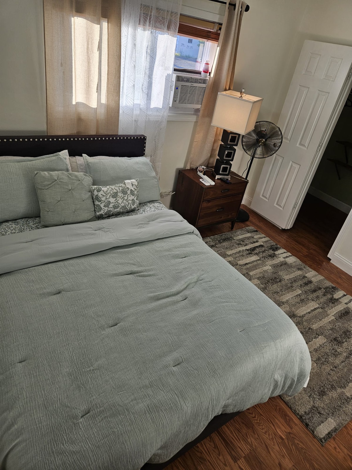 A neatly made king-size bed with soft gray bedding is centered in the room. Natural light enters through sheer curtains, illuminating the space. A bedside lamp and standing fan are positioned next to the bed, with a textured rug beneath. A closet is visible in the background.