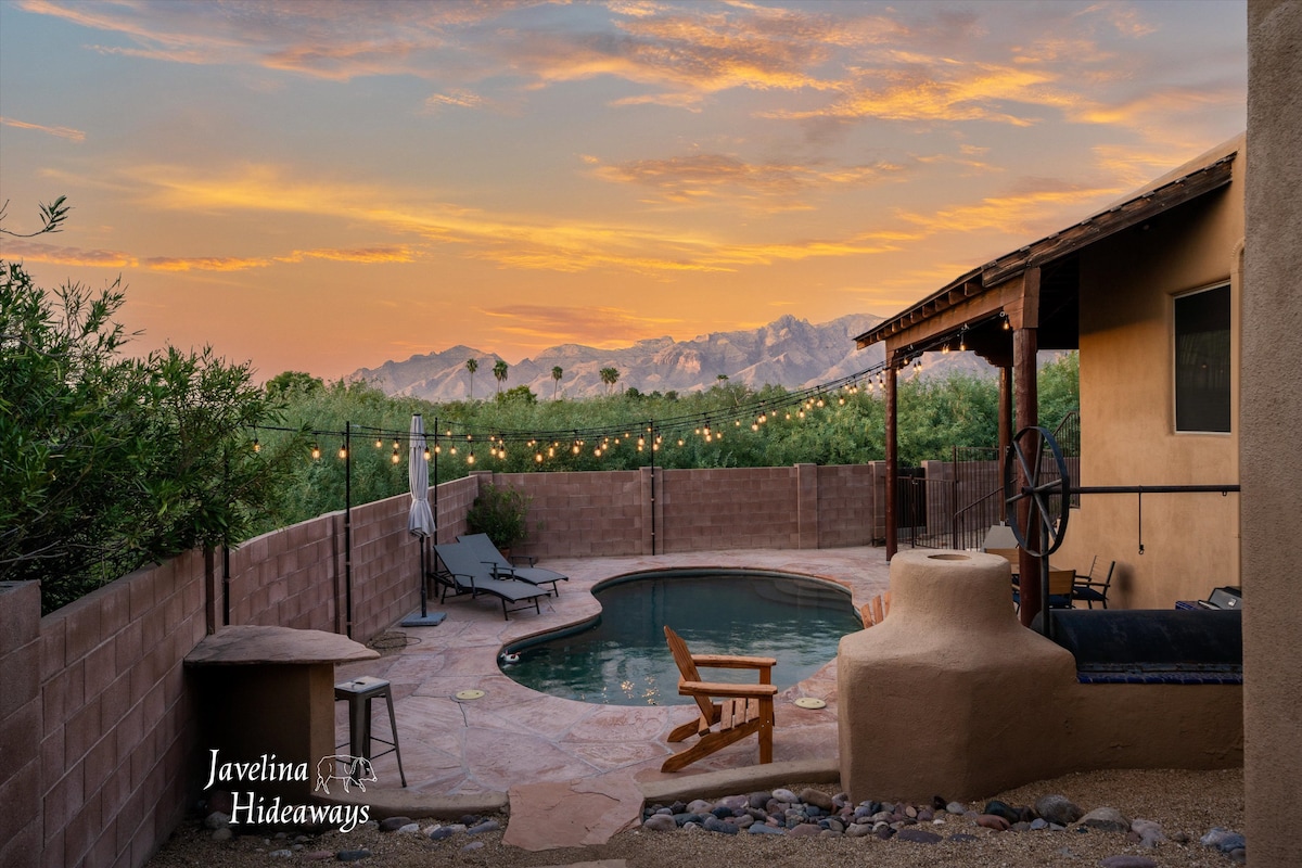 A serene outdoor space features a private pool surrounded by a patio, with comfortable lounge chairs and an inviting seating area. The backdrop includes a fence and lush greenery, while a warm sunset casts a soft glow over the scene, highlighting the mountain range in the distance.