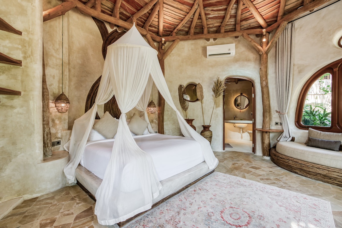 A circular room features a spacious bed with a light canopy, complemented by natural wood accents and intricate lighting fixtures. Large windows allow for abundant natural light, while a cozy seating area and elegant decor create a serene and inviting environment.