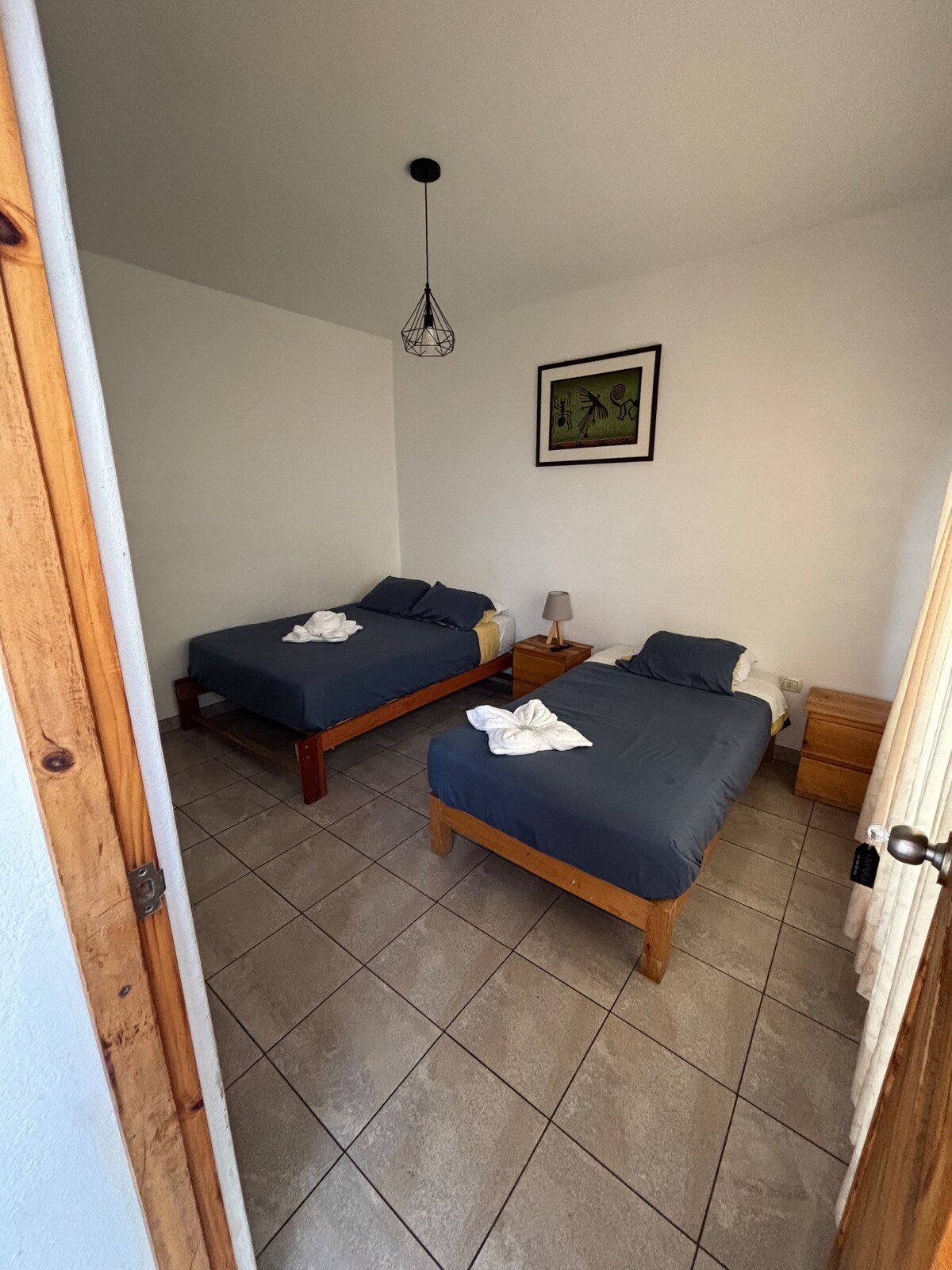 A simple bedroom is displayed with two beds positioned against the walls. Neutral wall colors create a calm environment. A small bedside table with a lamp is located next to one bed. A piece of artwork is hung above the furthest bed, and tiled flooring is present.