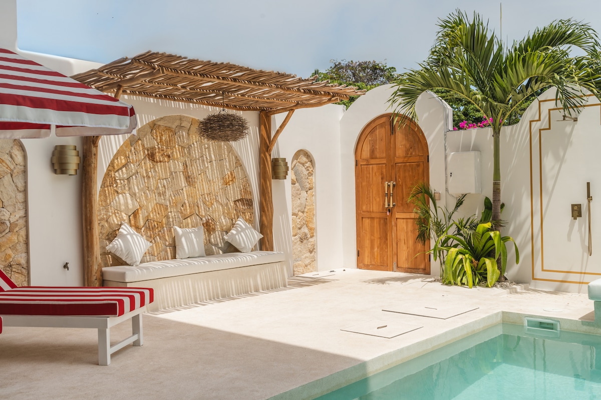 A sunlit outdoor area features a private pool surrounded by a spacious deck. A shaded, textured seating area with decorative cushions is set against a stone wall. Lush greenery adds a touch of nature, while a wooden door enhances the villa's charm.