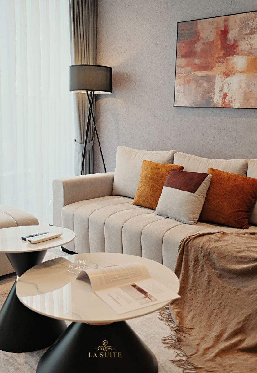 A modern living area features a light-colored sofa adorned with plush cushions in warm tones. Two minimalist side tables with glossy tops are positioned beside the sofa, complementing the contemporary decor. A soft throw blanket and an art piece with subtle colors enhance the inviting atmosphere.