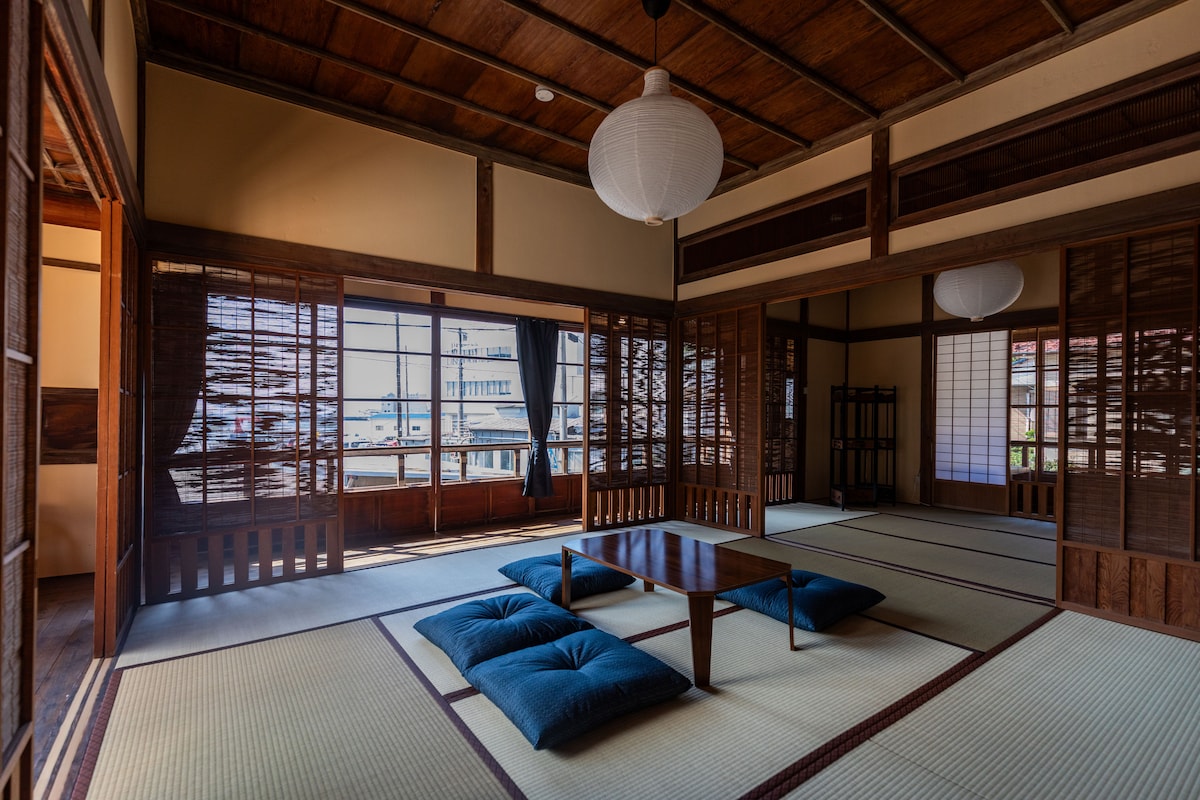 A spacious living area features tatami flooring and a low wooden table at the center. Cushions provide comfortable seating around the table. Shoji screens and large windows allow natural light to fill the room, offering views of the surroundings.