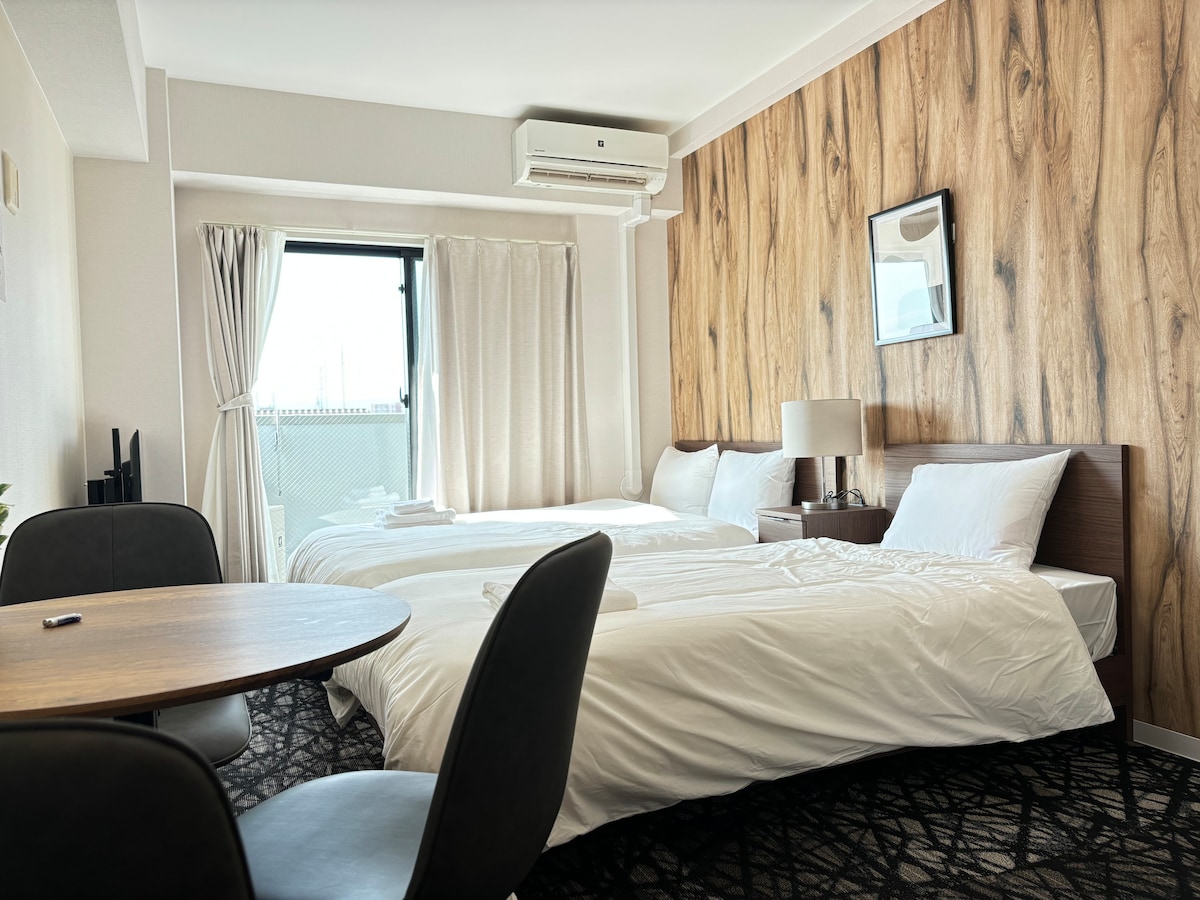 The room features two spacious beds arranged side by side, accompanied by a small round table with a chair. A window allows natural light to stream in, and the wall behind the beds displays a warm wood texture. An air conditioning unit is mounted above.