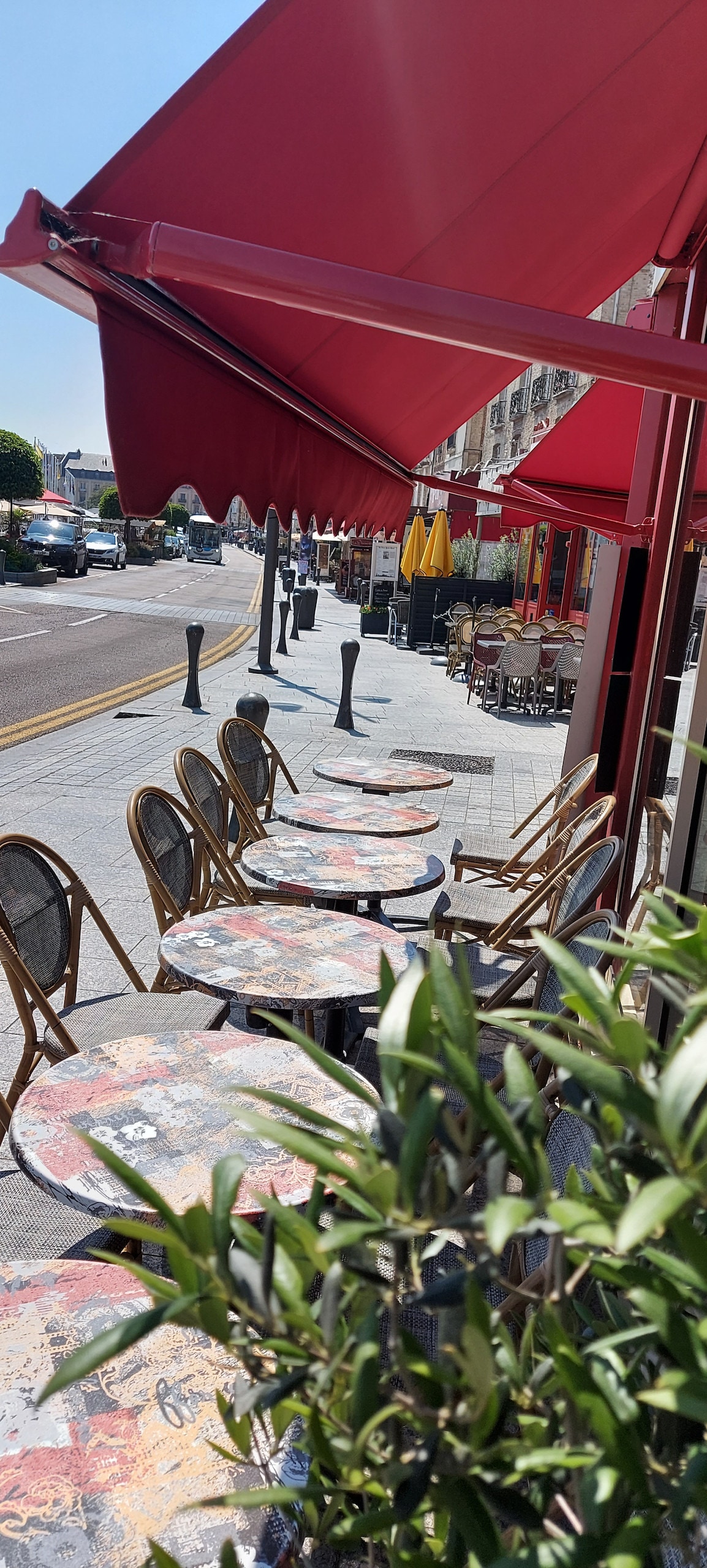 Several outdoor tables with patterned surfaces are arranged along a sunlit sidewalk, shaded by large red awnings. Bistro-style chairs complement the tables. The street features parked cars and distant greenery, suggesting a lively atmosphere near cafes and restaurants.
