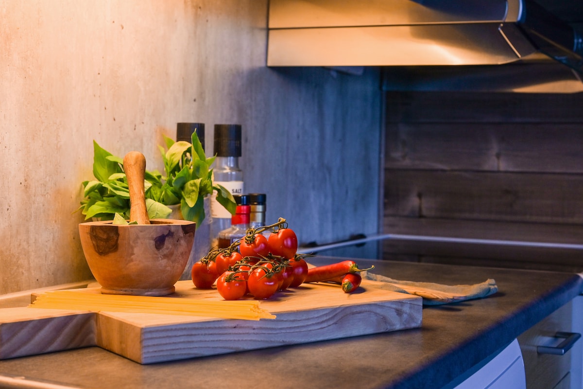 A kitchen countertop is displayed with a wooden bowl containing fresh basil, complemented by a mortar and pestle. Cherry tomatoes and red chili peppers are arranged on a wooden cutting board, with a sleek, modern backdrop featuring various kitchen utensils and an overhead cooktop.