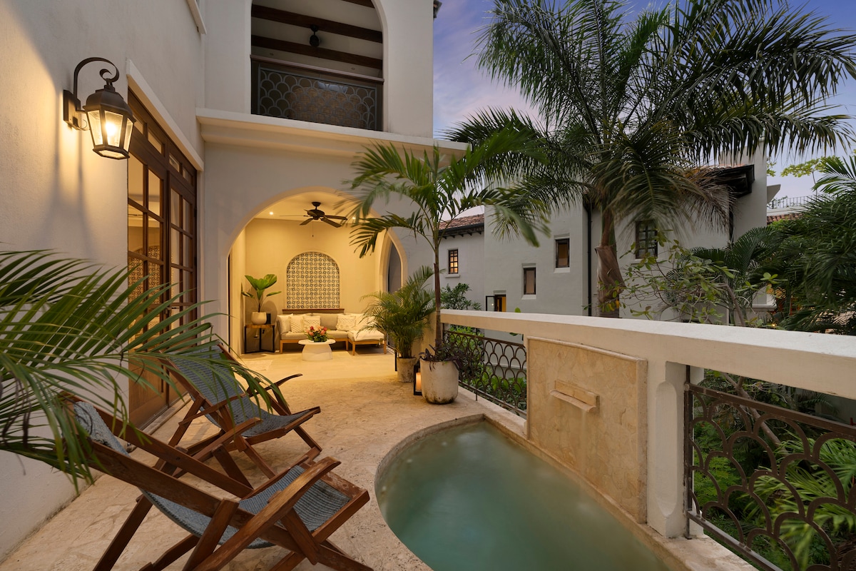 A covered patio area is showcased with comfortable seating and lush greenery surrounding a small plunge pool. Warm light from wall sconces creates a welcoming ambiance, while large windows offer views of the tranquil outdoor space and palm trees.