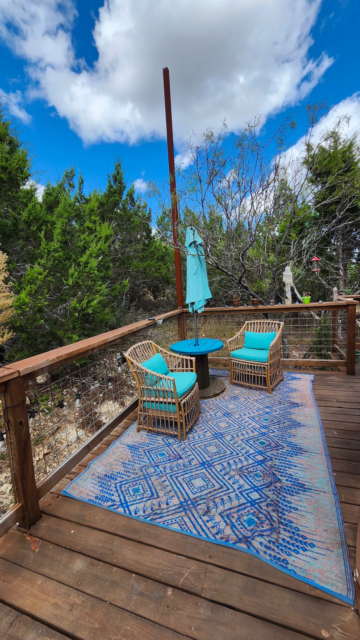 A spacious deck area is presented with two rattan chairs featuring blue cushions arranged around a small circular table. A vibrant blue rug anchors the seating arrangement, while a large umbrella provides shade. Lush greenery surrounds the space, enhancing the natural setting.
