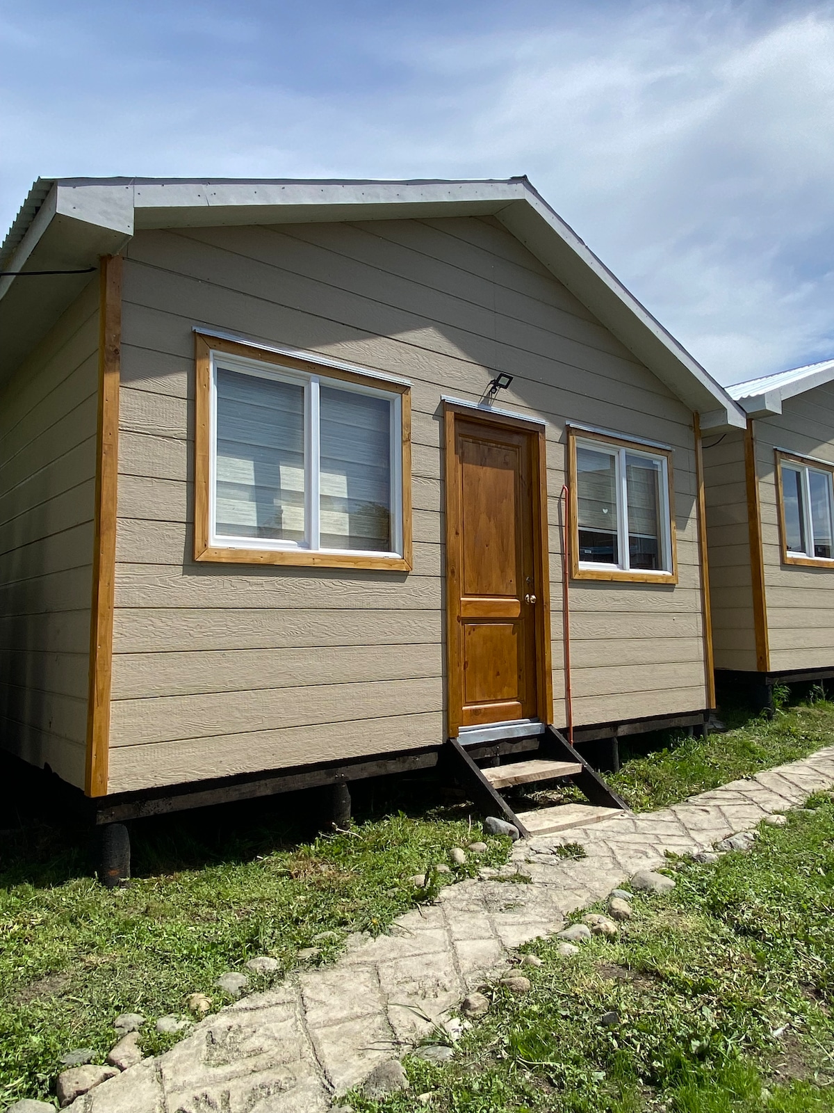 The exterior of a cabin is depicted, featuring light brown siding and a wooden door. Two large windows allow natural light to enter. A small staircase leads to the entrance, and a stone pathway is visible, bordered by greenery.